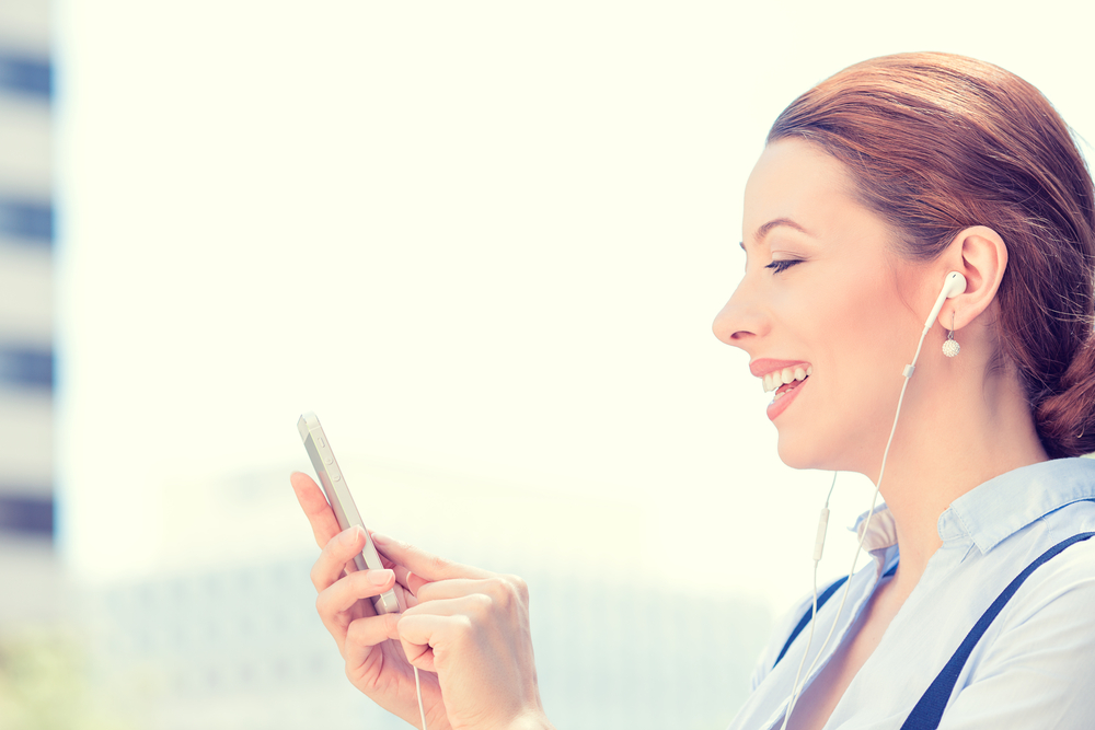 Closeup side view woman with earphones holding using smart mobile phone isolated outside corporate building background. People new generation technology concept. Customer service provider relationship